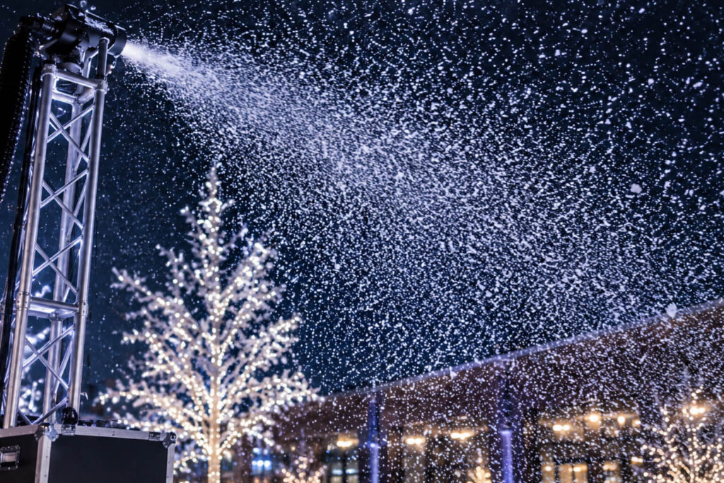 Close-up of a snow machine mounted on a metal truss blowing artificial snow into the night sky, illuminated by string-lit winter trees and outdoor festival lights in the background.