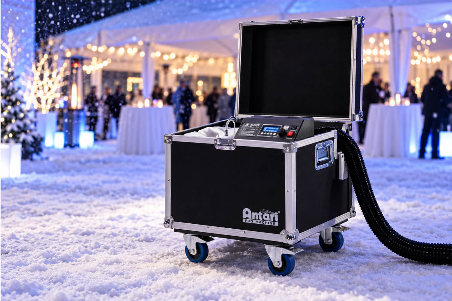 A black Antari snow machine in a road case sitting outdoors on snow, in front of a brightly lit event tent with festive holiday lights.